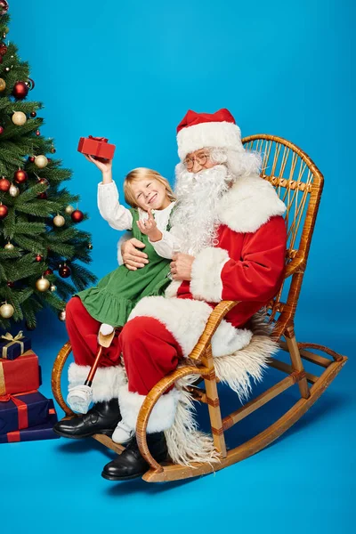 Ragazza felice con gamba protesica seduta sulle ginocchia di Babbo Natale con scatola regalo accanto all'albero di Natale — Foto stock