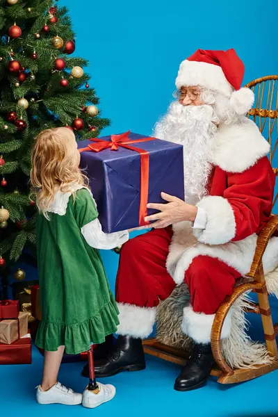 Санта Клаус дарит подарок счастливой девушке с протезной ногой рядом с елкой на синем фоне — стоковое фото