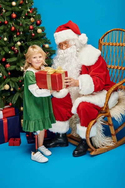 Santa dando regalo a la chica feliz con la pierna protésica al lado del árbol de Navidad en el fondo azul - foto de stock