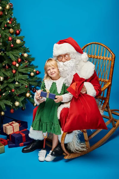 Fröhliches Mädchen mit Beinprothese hält Geschenk neben Weihnachtsmann neben Weihnachtsbaum auf blau — Stockfoto