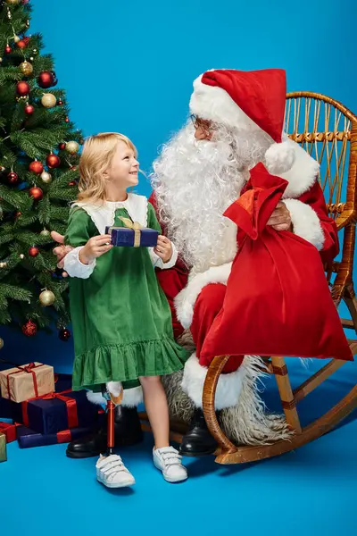 Счастливая девочка с протезом ноги держит подарок рядом с елкой на голубом фоне — стоковое фото