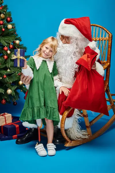 Alegre chica con prótesis pierna celebración presente cerca de Santa Claus junto a árbol de Navidad en azul - foto de stock