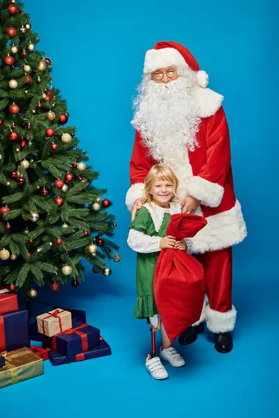 Счастливая девушка с протезной ногой держит мешок рядом с Санта-Клаусом рядом с рождественской елкой на голубом — стоковое фото
