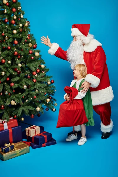 Alegre chica con prótesis pierna celebración saco cerca de Santa Claus al lado del árbol de Navidad en azul - foto de stock