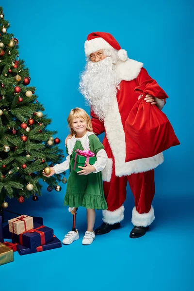 Santa Claus holding sack bag near cheerful girl with prosthetic leg next to Christmas tree on blue — Stock Photo