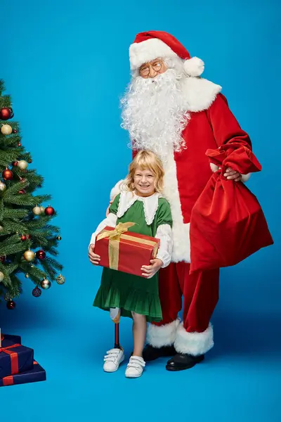 Санта Клаус держит мешок с мешком рядом с веселым ребенком с протезной ногой рядом с рождественской ёлкой на синей — стоковое фото