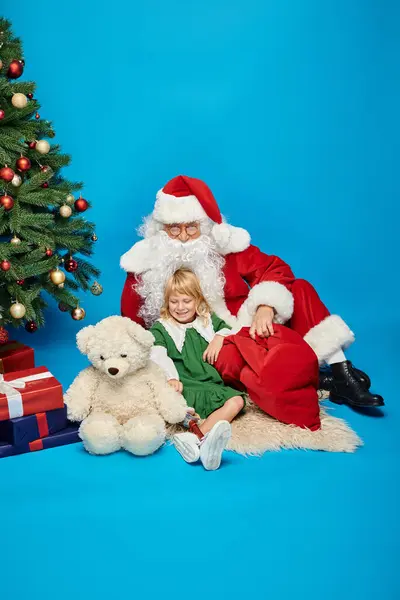 Chica feliz con la pierna protésica y el oso de peluche y sentado con Santa Claus al lado del árbol de Navidad - foto de stock