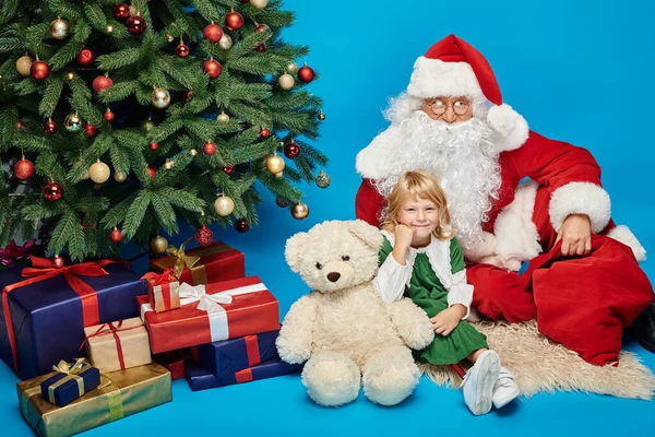 Niño feliz con la pierna protésica y el oso de peluche sentado con Santa Claus al lado del árbol de Navidad - foto de stock