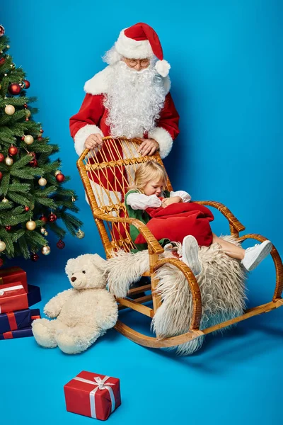 Santa Claus mecedora con linda chica con prótesis pierna bolsa de sujeción cerca del árbol de Navidad - foto de stock