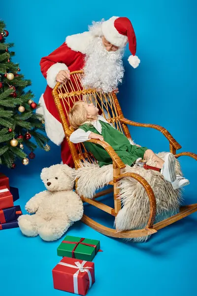 Santa Claus mecedora con linda chica con pierna protésica al lado de oso de peluche y árbol de Navidad - foto de stock