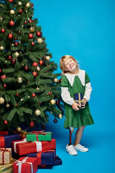 Niña alegre con la pierna protésica sosteniendo regalo envuelto al lado del árbol de Navidad en azul - foto de stock