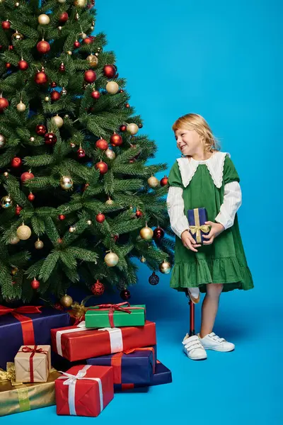 Joyful little girl with prosthetic leg holding wrapped present next to Christmas tree on blue — Stock Photo