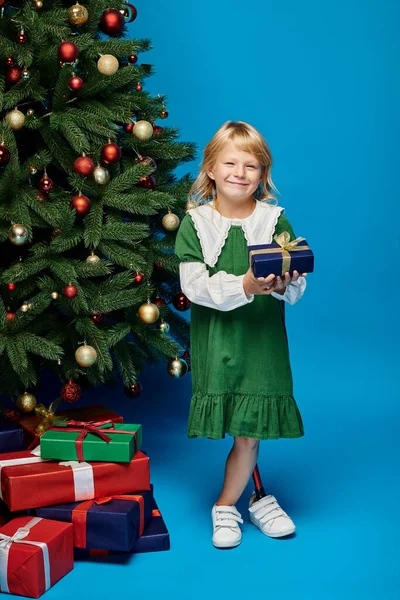 Счастливая девушка в платье с протезной ногой держа завернутый подарок рядом с елкой на голубой — стоковое фото