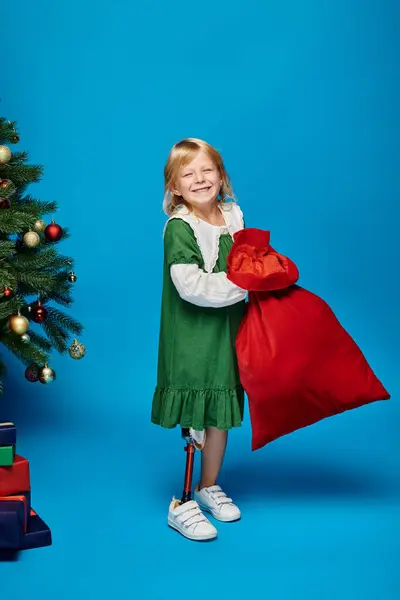 Chica feliz en vestido con prótesis de la pierna bolsa de sujeción con regalos cerca del árbol de Navidad en azul - foto de stock