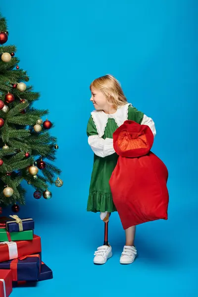 Chica complacida en vestido con prótesis de pierna bolsa de sujeción con regalos cerca del árbol de Navidad en azul - foto de stock