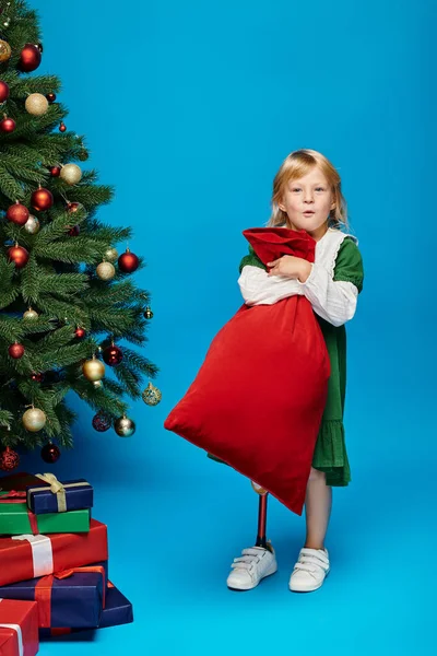 Amazed girl in dress with prosthetic leg holding sack bag with presents near Christmas tree on blue — Stock Photo
