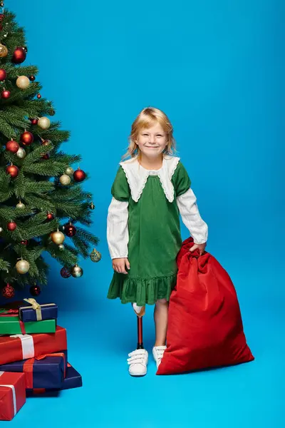 Довольный ребенок в платье с протезной ногой держа мешок с подарками возле елки на голубой — стоковое фото