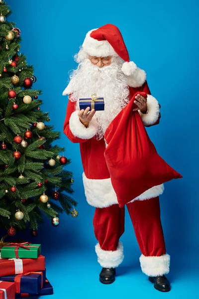 Feliz Papá Noel con barba y anteojos sosteniendo bolsa de saco y presente cerca del árbol de Navidad - foto de stock