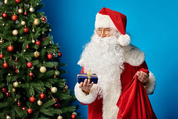 Happy Santa Claus with beard and eyeglasses in red costume holding sack bag and Christmas present — Stock Photo