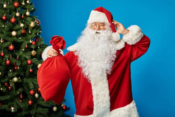 Emocionado Santa Claus con barba y gafas que sostienen el saco con regalos de Navidad en azul - foto de stock
