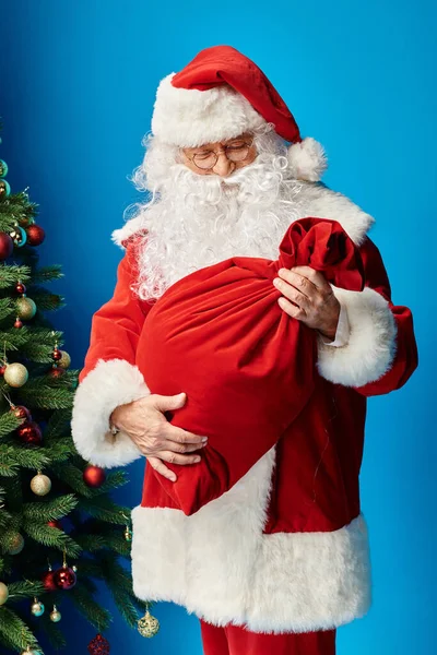 Santa Claus with beard and eyeglasses looking at red sack bag with Christmas presents on blue — Stock Photo