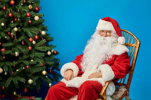 Joyeux Père Noël avec barbe et lunettes assis dans un fauteuil à bascule près de l'arbre de Noël sur bleu — Photo de stock