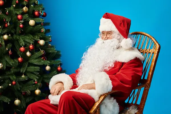 Щасливий Санта Клаус з бородою і окулярами, сидячи в кріслі-качалці біля ялинки на синьому — стокове фото