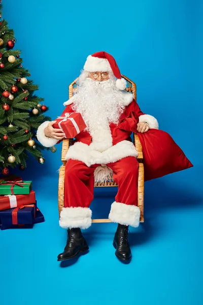 Feliz Santa Claus en gafas de vista sentado en mecedora con regalo y bolsa cerca del árbol de Navidad - foto de stock