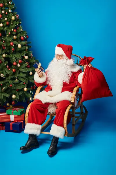 Glücklicher Weihnachtsmann im Schaukelstuhl sitzend mit Geschenk und Sacksack neben Weihnachtsbaum auf blau — Stockfoto