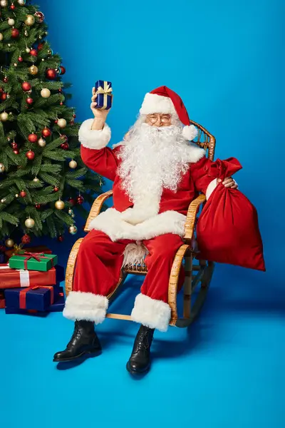 Alegre Papai Noel sentado em cadeira de balanço com presente e saco saco perto da árvore de Natal em azul — Fotografia de Stock