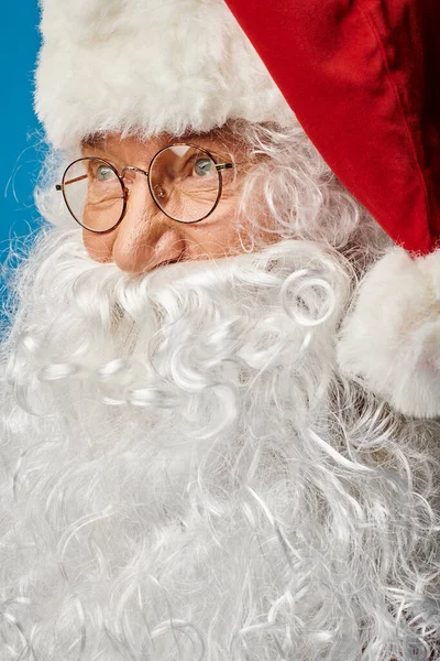 Primer plano de Santa Claus feliz con barba blanca y anteojos mirando hacia otro lado sobre fondo azul - foto de stock