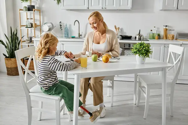 Feliz madre rubia mirando a su linda hija con prótesis pierna desayunando en la cocina - foto de stock