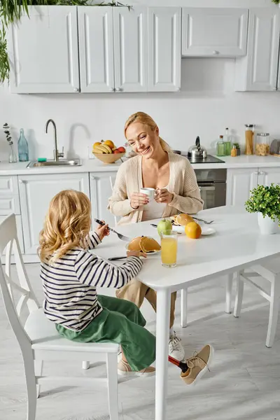 Улыбающаяся мать-блондинка смотрит на свою милую дочь с протезной ногой, завтракающую на кухне — стоковое фото