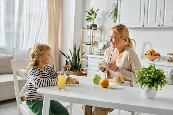 Cheerful blonde mother looking at her cute daughter with prosthetic leg having breakfast in kitchen — Stock Photo