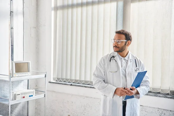 Jovem médico indiano com óculos e estetoscópio em enfermaria hospitalar olhando para longe, cuidados de saúde — Fotografia de Stock