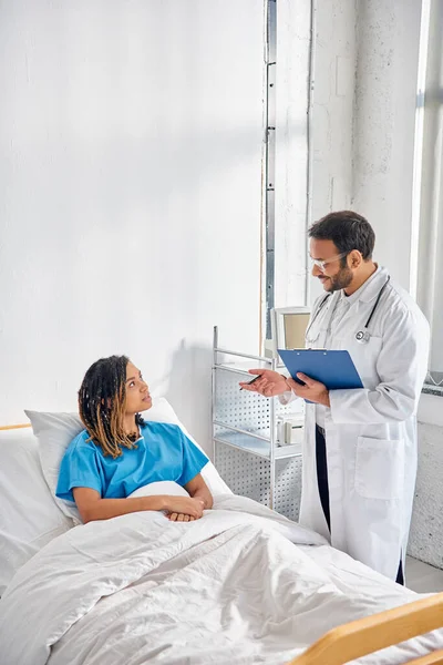 Young indian doctor looking at african american patient lying in bed in hospital ward, healthcare — Stock Photo