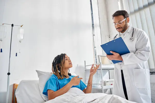 Giovane medico indiano guardando il paziente afro-americano sdraiato nel letto nel reparto ospedaliero, assistenza sanitaria — Foto stock