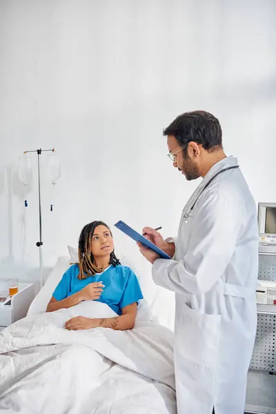 Attraente donna afro-americana sdraiata a letto nel reparto ospedaliero parlando con il suo giovane medico indiano — Foto stock
