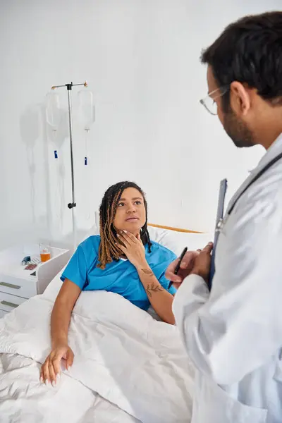 Attraente donna afro-americana sdraiata a letto nel reparto ospedaliero parlando con il suo giovane medico indiano — Foto stock