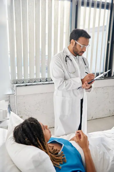 Médico indio bien parecido tomando notas sobre el paciente afroamericano acostado en la cama en la sala de hospital - foto de stock