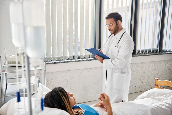 Good looking indian doctor taking notes on african american patient lying in bed in hospital ward — Stock Photo