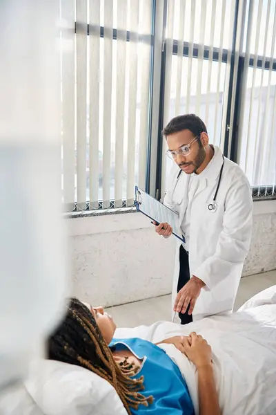 Doctora india inteligente hablando con su paciente afroamericana acostada en la cama en la sala de salud - foto de stock