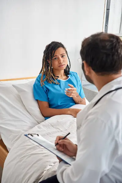 Atractiva mujer afroamericana enferma acostada en la cama de hospital hablando con su joven médico indio - foto de stock