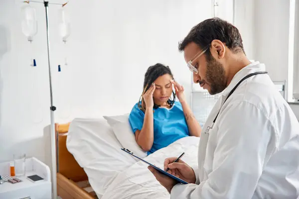 Focus on young indian doctor sitting on bed near his blurred african american patient with headache — Stock Photo