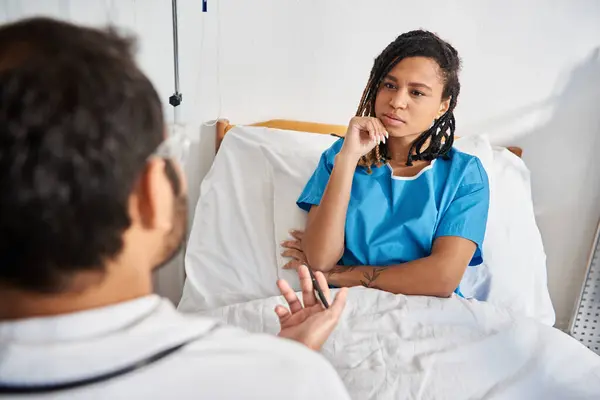 Attraktive kranke afrikanisch-amerikanische Frau liegt im Krankenhausbett und hört ihrem jungen indischen Arzt zu — Stockfoto