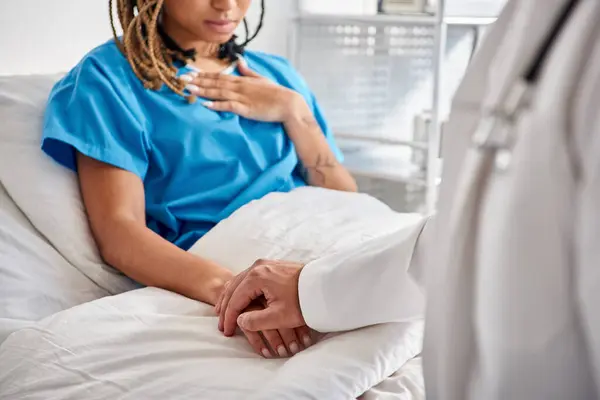 Visão recortada de jovem médico indiano segurando sua mão paciente afro-americana deitada na cama do hospital — Fotografia de Stock