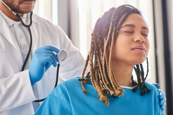 Médico indio escuchando el aliento de su paciente afroamericano enfermo con estetoscopio, atención médica - foto de stock