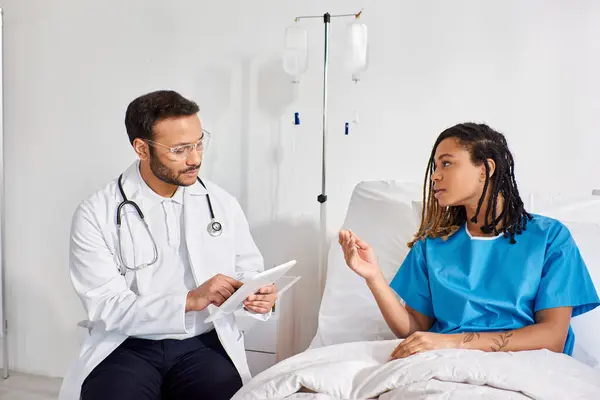 African american patient lying in hospital bed talking to her indian doctor taking notes on tablet — Stock Photo