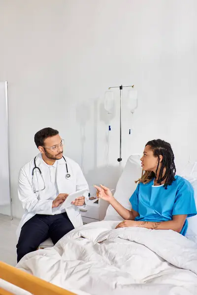 Paziente afroamericano sdraiato nel letto d'ospedale a parlare con il suo medico indiano prendendo appunti su tablet — Foto stock