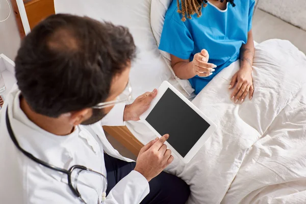 Paziente afroamericano seduto sul letto vicino al suo medico indiano prendendo appunti su tablet, ritagliato — Foto stock